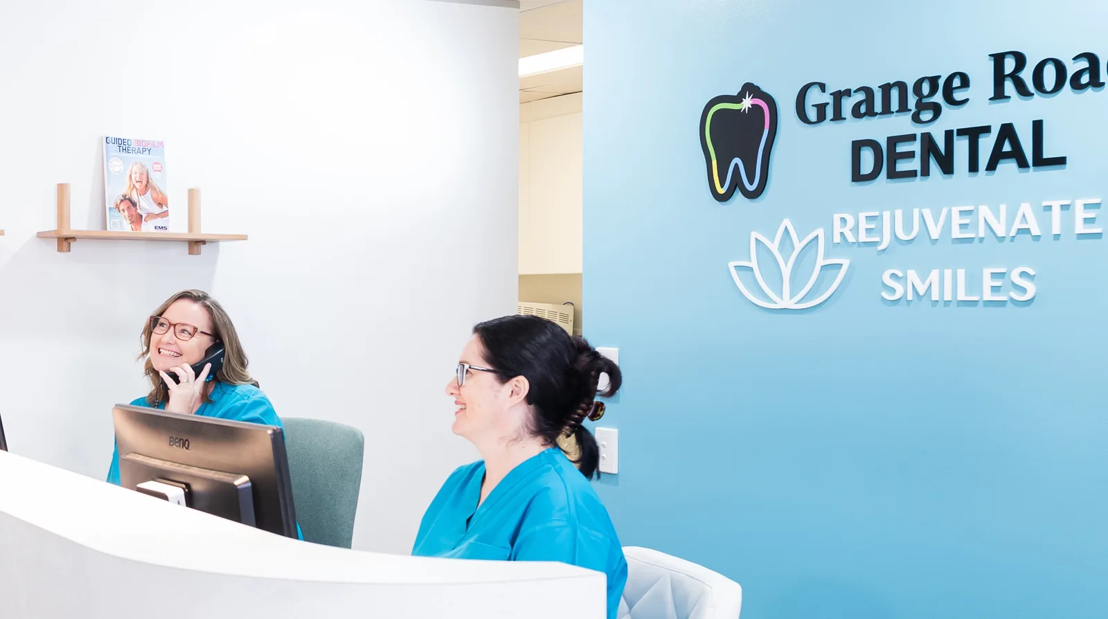 Two smiling members of our front desk team at the reception desk beside the teal Grange Road Dental wall in Ipswich, one of them chatting on the phone